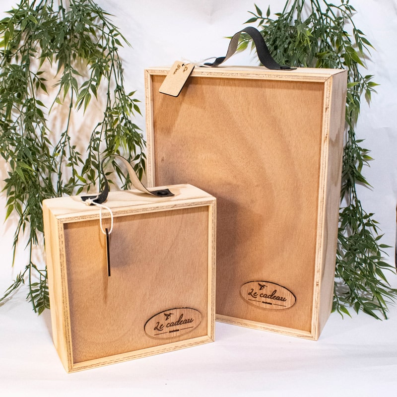 Dos cajas de madera fabricadas en tríplex de pino, con un diseño artesanal y elegante. Ambas tienen tapa deslizable, destacándose sus acabados lisos que permiten apreciar las vetas naturales del material. La caja más pequeña es cuadrada, mientras que la más grande tiene forma rectangular alargada. En la parte inferior de cada caja, un grabado láser personalizado muestra el texto 