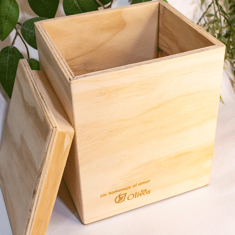 Una elegante caja de madera cuadrada con tapa, elaborada en un tono cálido y natural. Presenta un diseño sencillo y sofisticado, con líneas rectas y una tapa que encaja perfectamente. En la parte frontal de la caja se observa un grabado delicado con la leyenda 