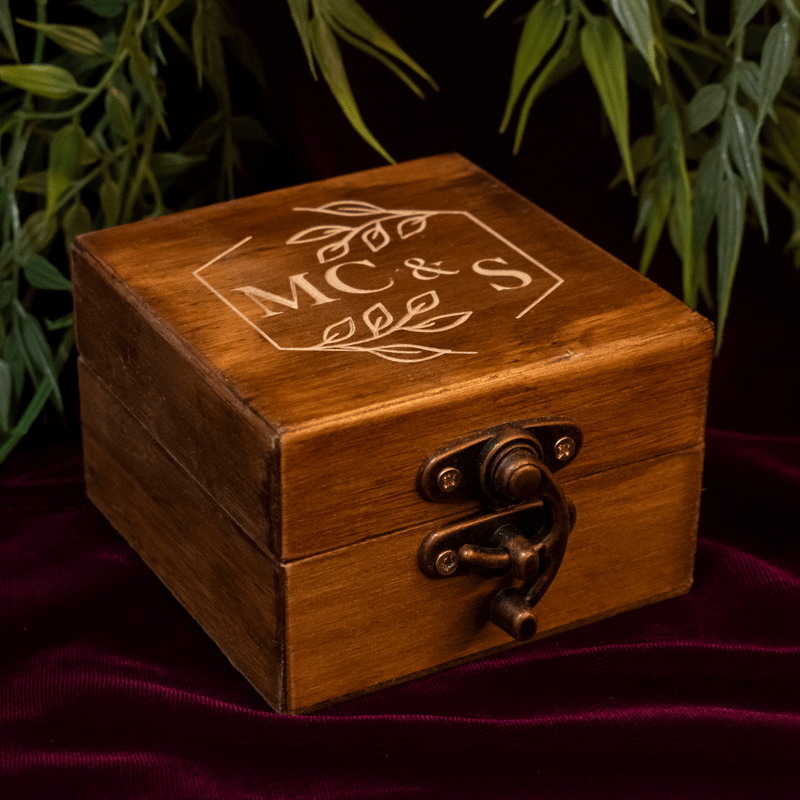 Elegante caja para anillos elaborada en madera de alta calidad, con un diseño rústico y sofisticado. Su forma cuadrada y compacta, junto con el cierre de metal envejecido, le confieren un aire vintage y romántico. La tapa de la caja está personalizada con un grabado láser que puede incluir las iniciales de los novios, la fecha de la boda, un mensaje especial o un diseño personalizado, como se muestra en la imagen con las iniciales 