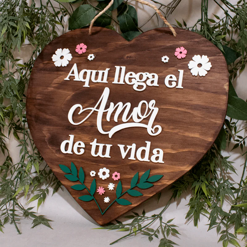 Hermoso letrero en forma de corazón, elaborado en madera natural con un acabado rústico y romántico. El corazón está decorado con la frase "Aquí llega el Amor de tu vida" escrita en una elegante tipografía y acompañada de delicadas flores y ramas pintadas a mano. El letrero cuenta con una cuerda de yute para colgarlo fácilmente en cualquier lugar. Es el complemento perfecto para decorar la ceremonia de boda o el hogar de los recién casados.